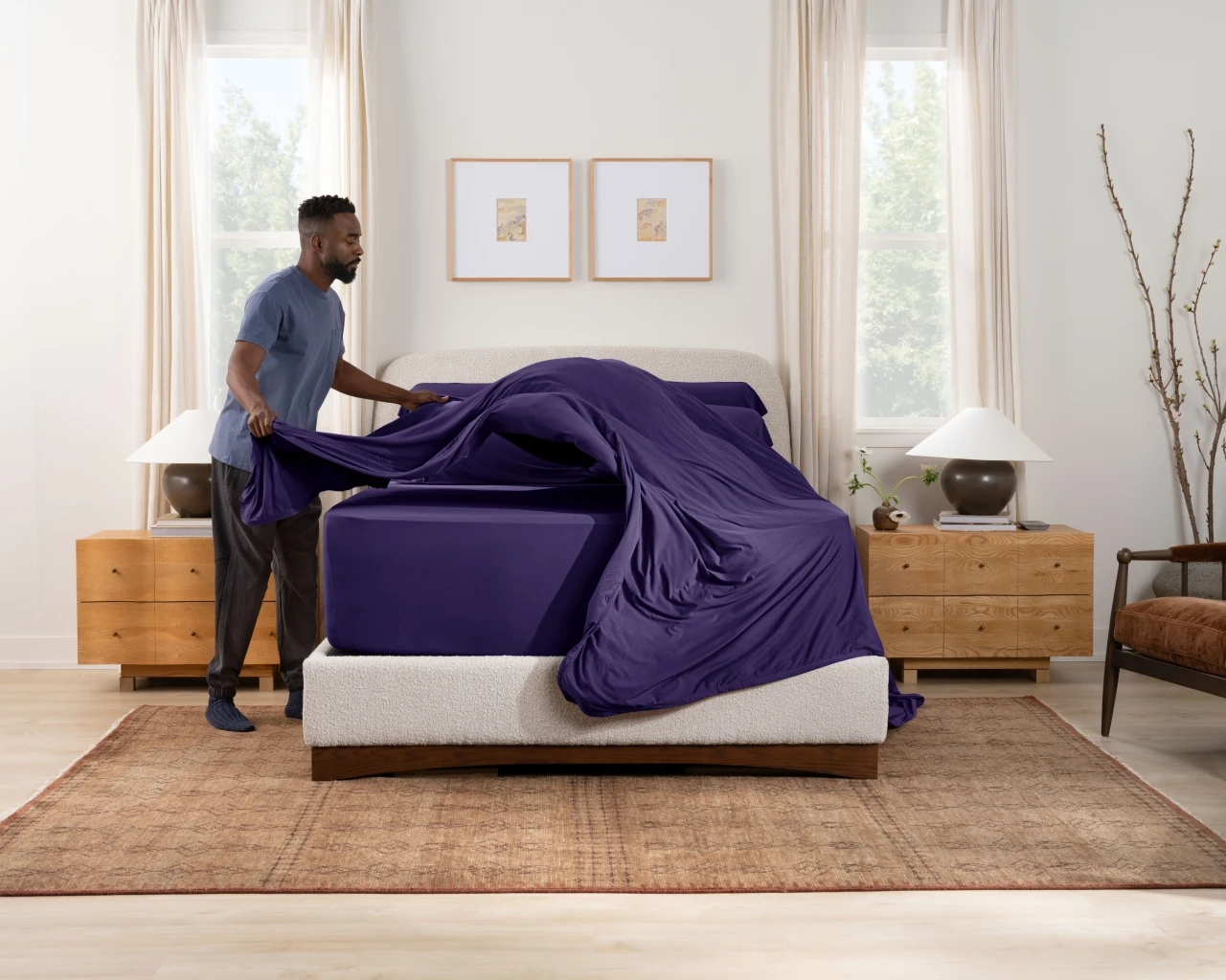 A man is making the bed, pulling up a purple sheet. The furniture around the bed is simple, and sunlight streams into the room through the curtains, creating a fresh and natural atmosphere.