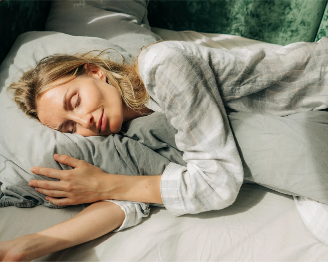A woman lies on her side in bed, hugging a pillow, with a smile on her face, looking peaceful and comfortable, against a warm background.