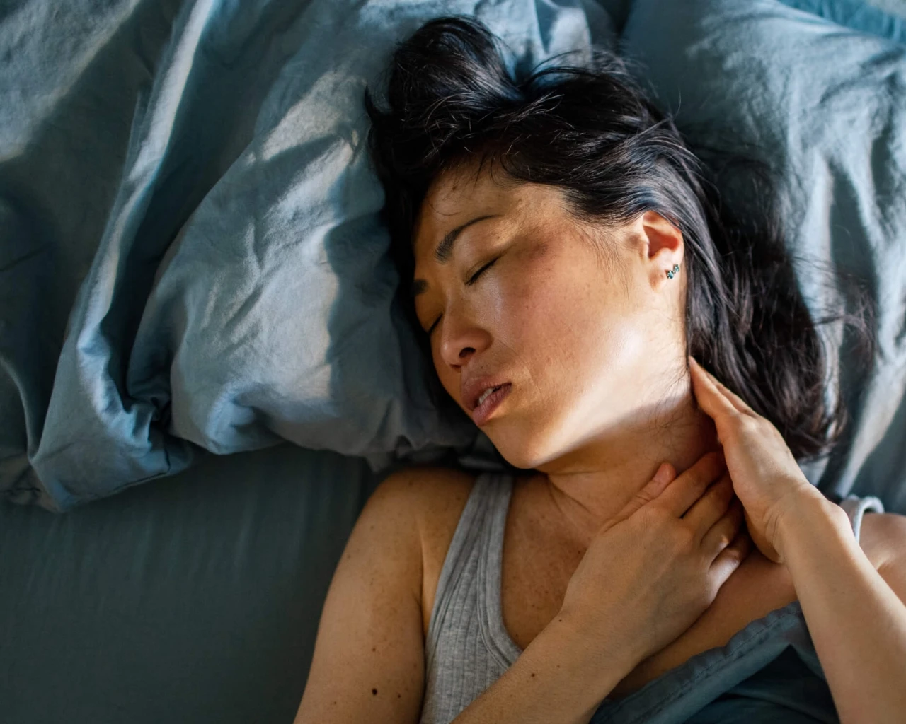 A woman lies in bed, her brow furrowed as if she has a neck ache. The bed is covered with blue sheets and pillows, and the environment is serene.