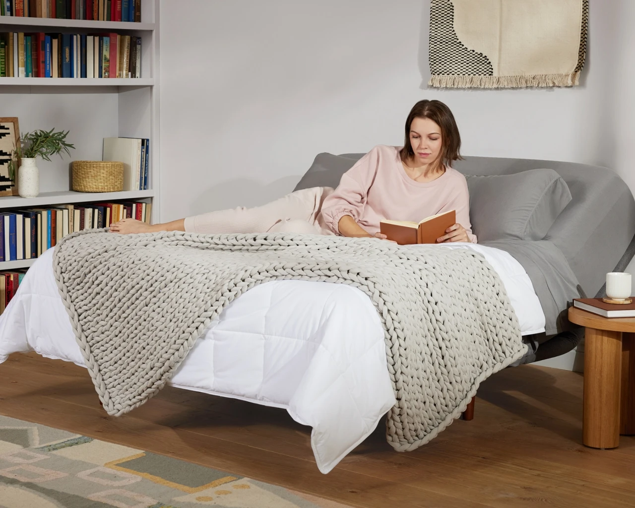 A woman wearing comfortable pink pajamas sits on a bed reading a book. The bed is covered with simple white sheets and a gray blanket. The surrounding environment is warm and peaceful, with a bookshelf in the background.