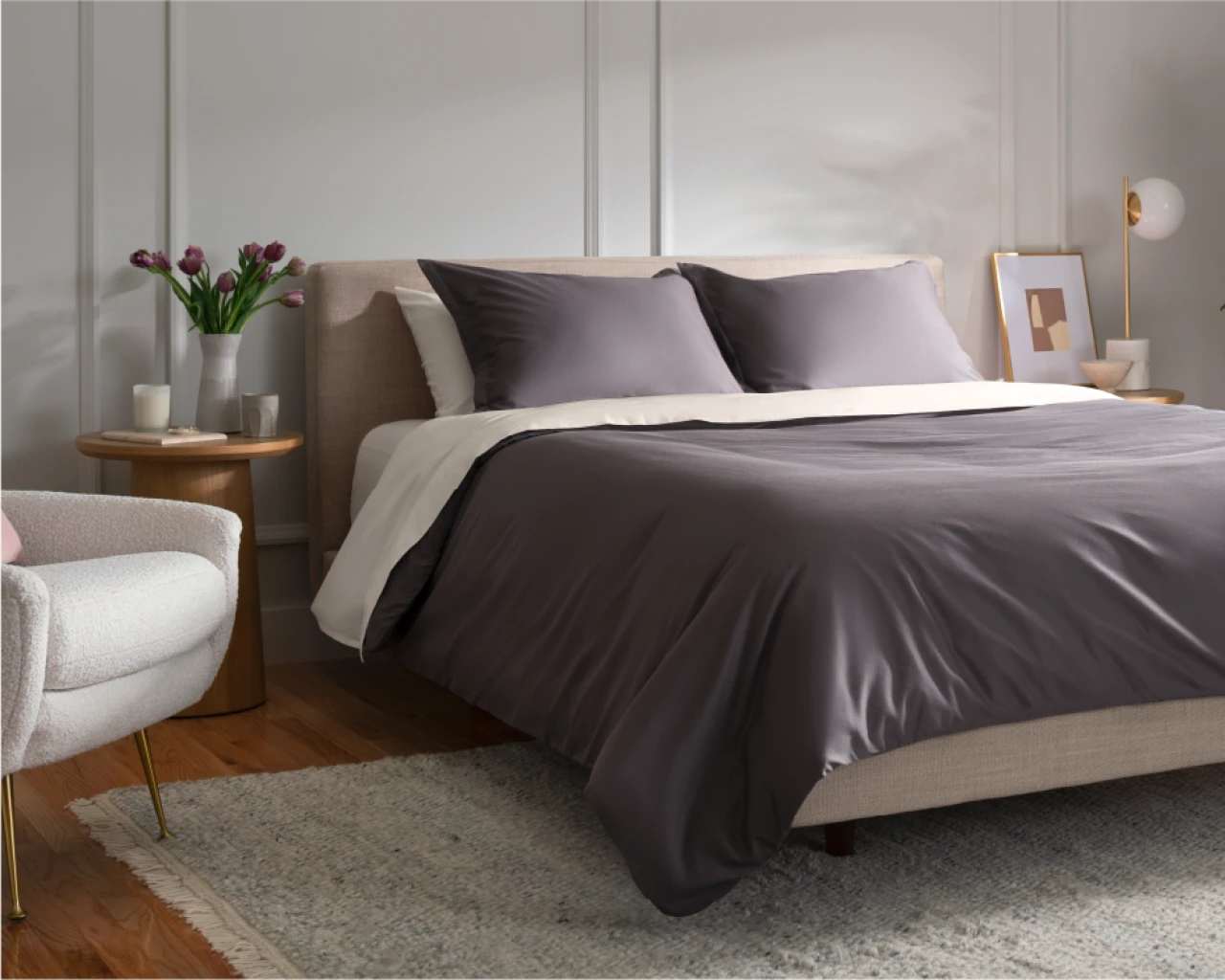 A modern bedroom with dark gray sheets and pillows on the bed, a vase of tulips and a decorative painting on the side; the overall environment is warm and simple.