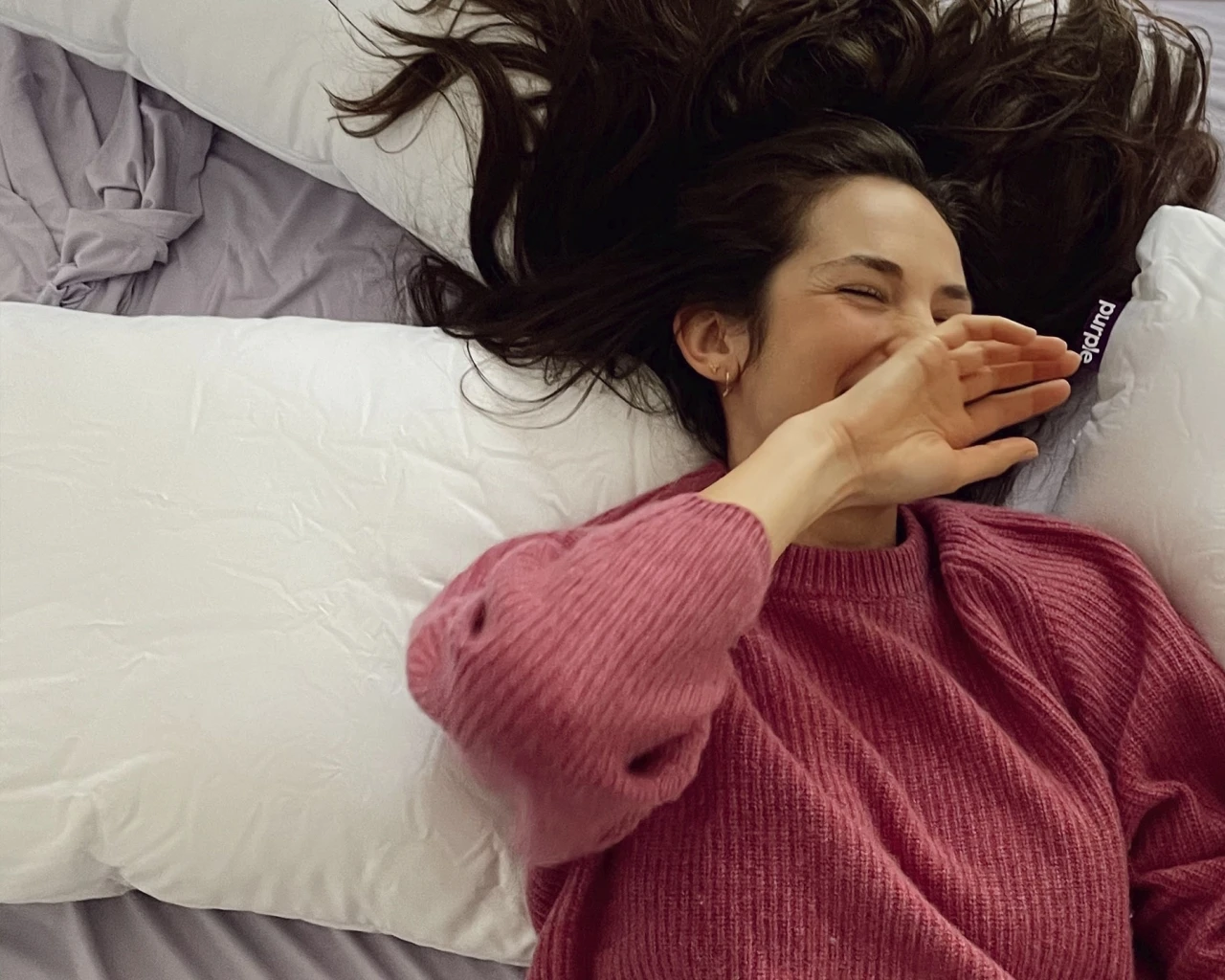 A woman lies on the bed with her hair down, wearing a pink sweater, smiling and covering her mouth. The scene is relaxed and pleasant, with light-colored pillows and sheets on the bed.