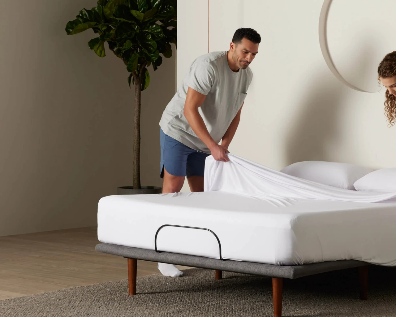 A couple is making the bed. One person is pulling up the sheets on the other side of the bed while the other person stands beside the bed. The whole scene presents a warm and comfortable bedroom atmosphere.