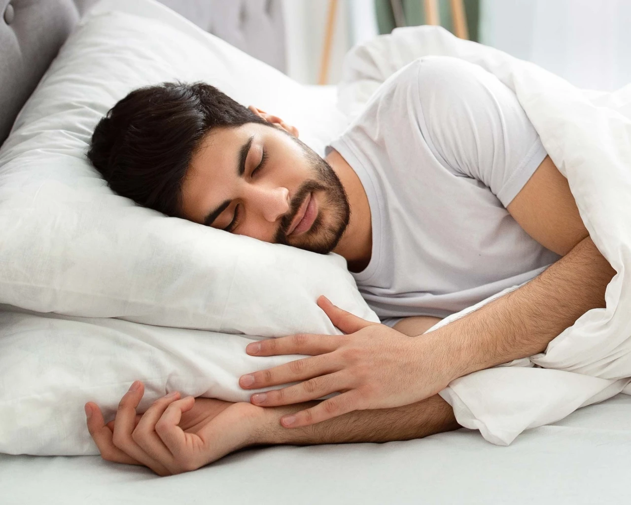 A man is fast asleep on his side, hugging a white pillow. He looks relaxed, and the scene is peaceful and comfortable.
