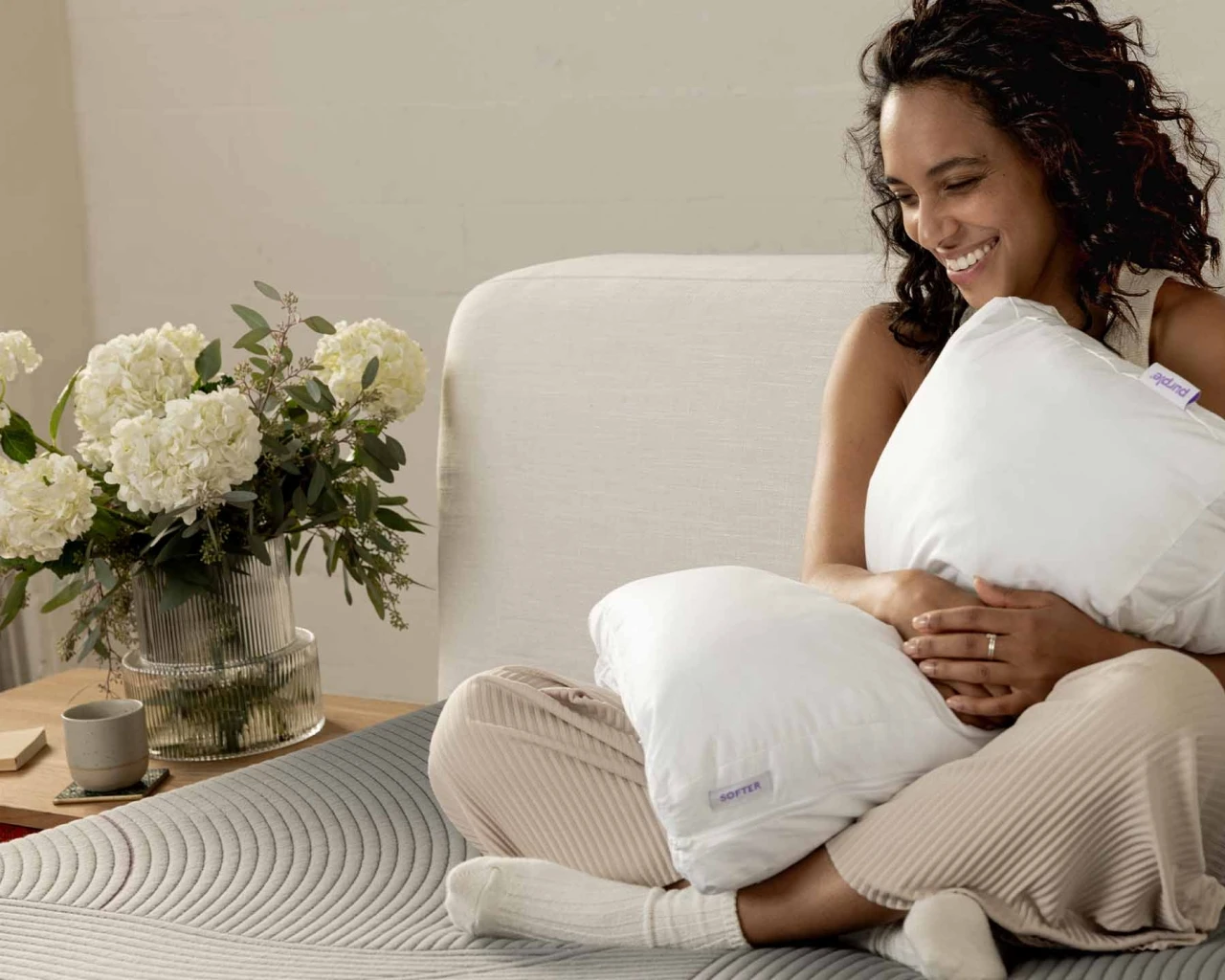 A woman sits on the bed, hugging two pillows and smiling. In the background, there is a vase of white flowers and a cup of tea, creating a warm and comfortable atmosphere.