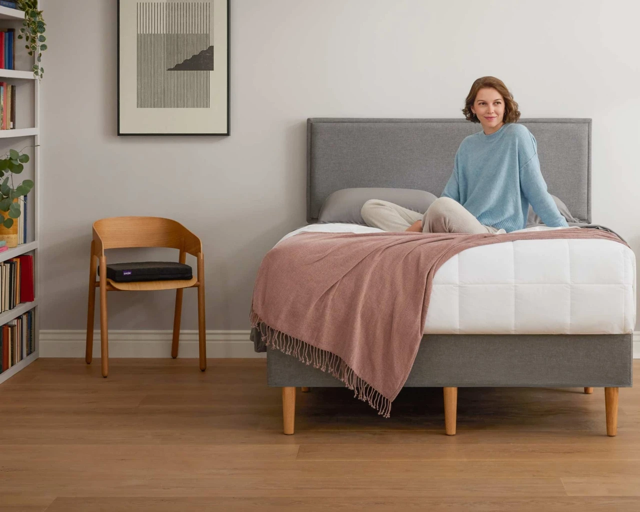 A woman wearing a blue sweater sits on a bed covered with a pink blanket. The background is a simple bedroom design, with a bookshelf and a wooden chair nearby, creating a fresh and cozy atmosphere.